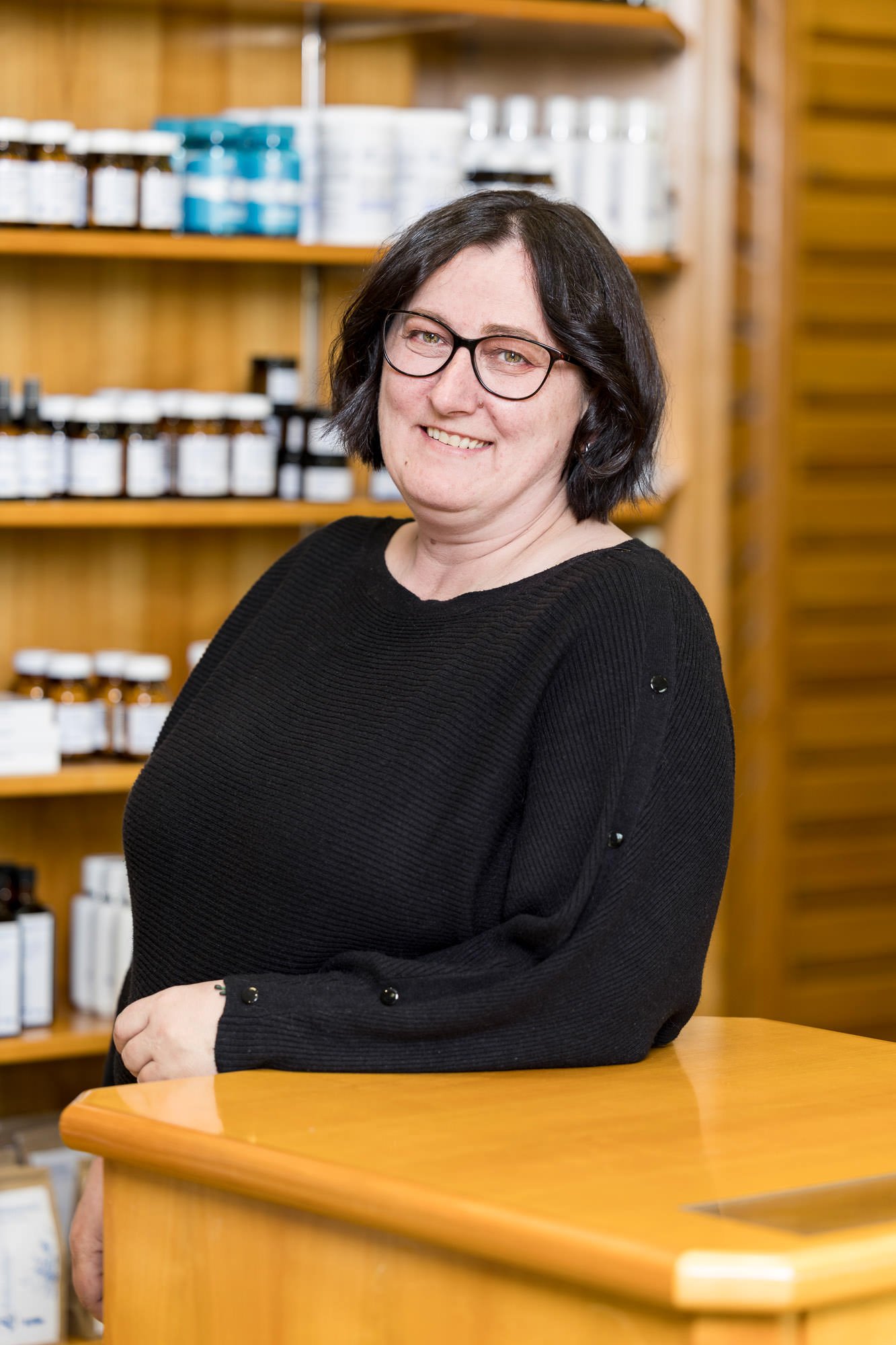 Porträt einer lächelnden Frau mit Brille, die in einem Geschäft für Naturprodukte steht. Sie trägt einen schwarzen Pullover und steht an einem Holztresen, im Hintergrund sind Regale mit Flaschen und Behältern sichtbar.