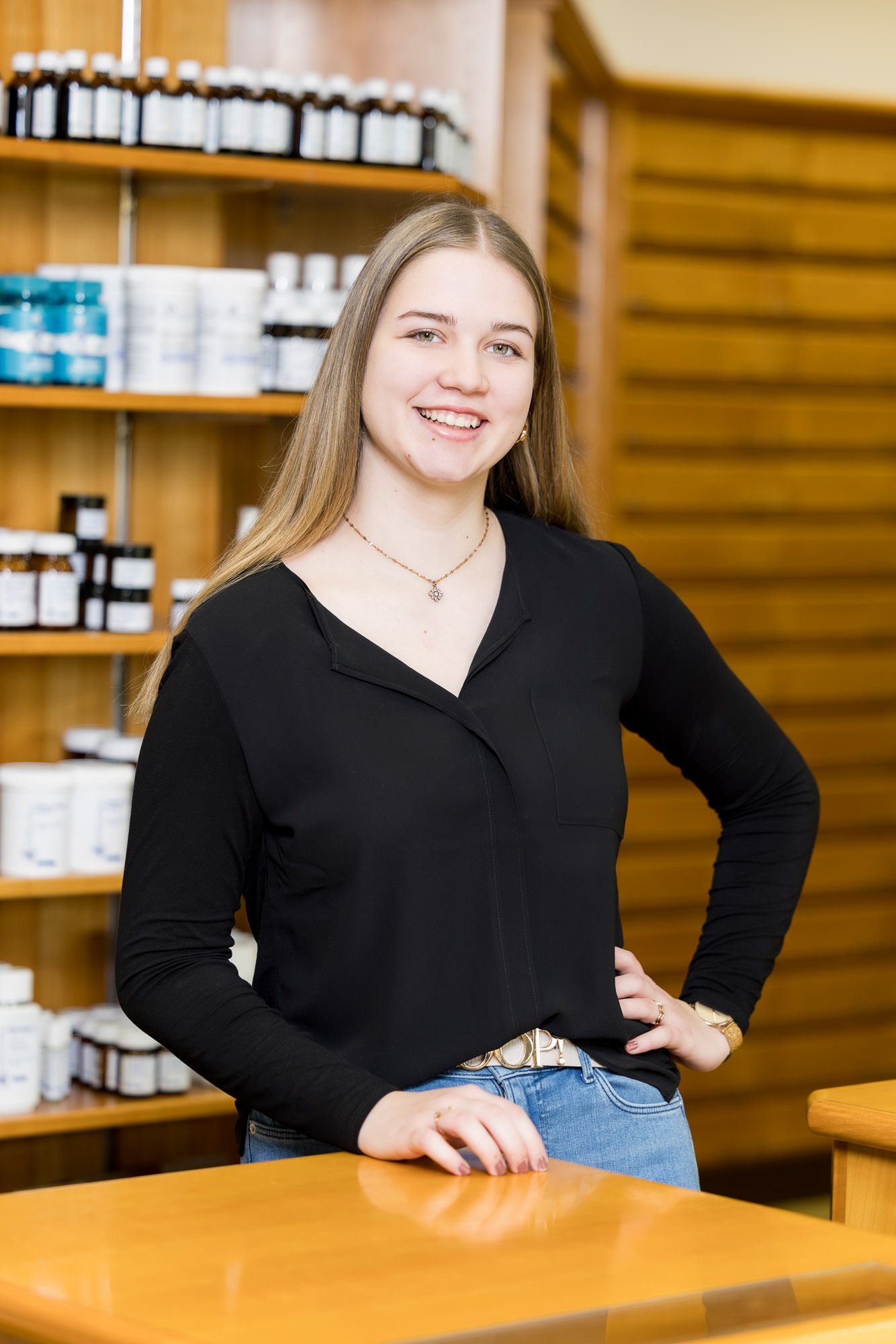 Junge Frau mit langen Haaren lächelt und steht an einem Holztisch in einem Geschäft, umgeben von Regalen mit verschiedenen Produkten. Sie trägt ein schwarzes Oberteil und Jeans, während sie mit einer Hand auf den Tisch gestützt ist.