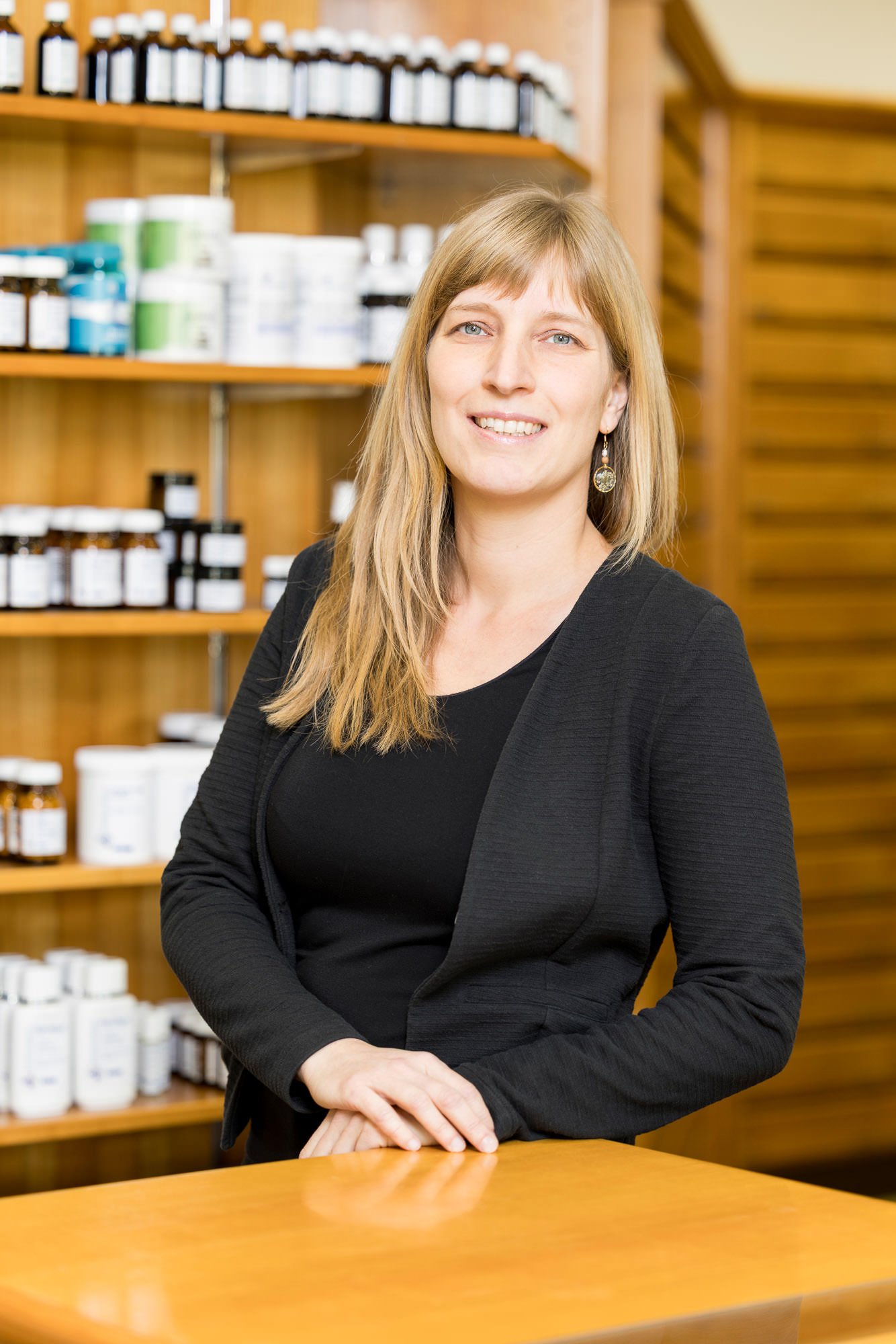 Eine Frau mit langen, blonden Haaren steht an einem Holztisch in einem Geschäft für Naturprodukte. Im Hintergrund sind Regale mit verschiedenen Flaschen und Behältern sichtbar. Die Frau trägt ein schwarzes Oberteil und hat einen freundlichen Gesichtsausdruck.
