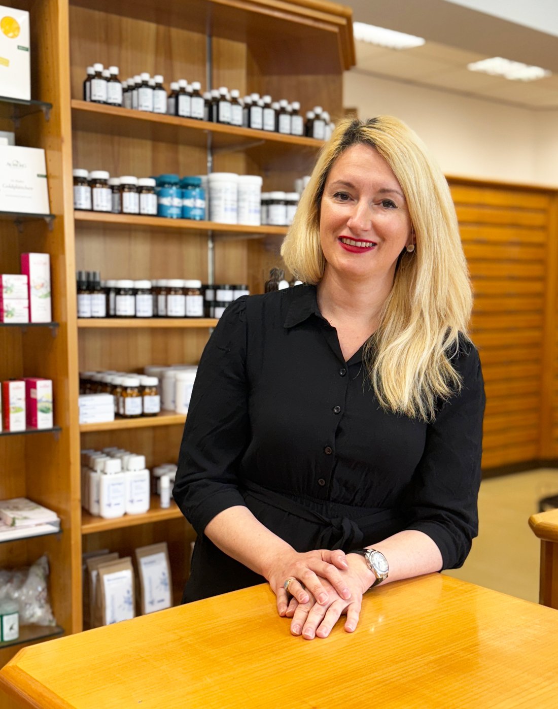 Eine blonde Frau in einem schwarzen Kleid steht an einem Holztisch in einem Geschäft für natürliche Produkte. Im Hintergrund sind Regale mit verschiedenen Flaschen und Verpackungen sichtbar. Die Frau lächelt und trägt eine Armbanduhr.