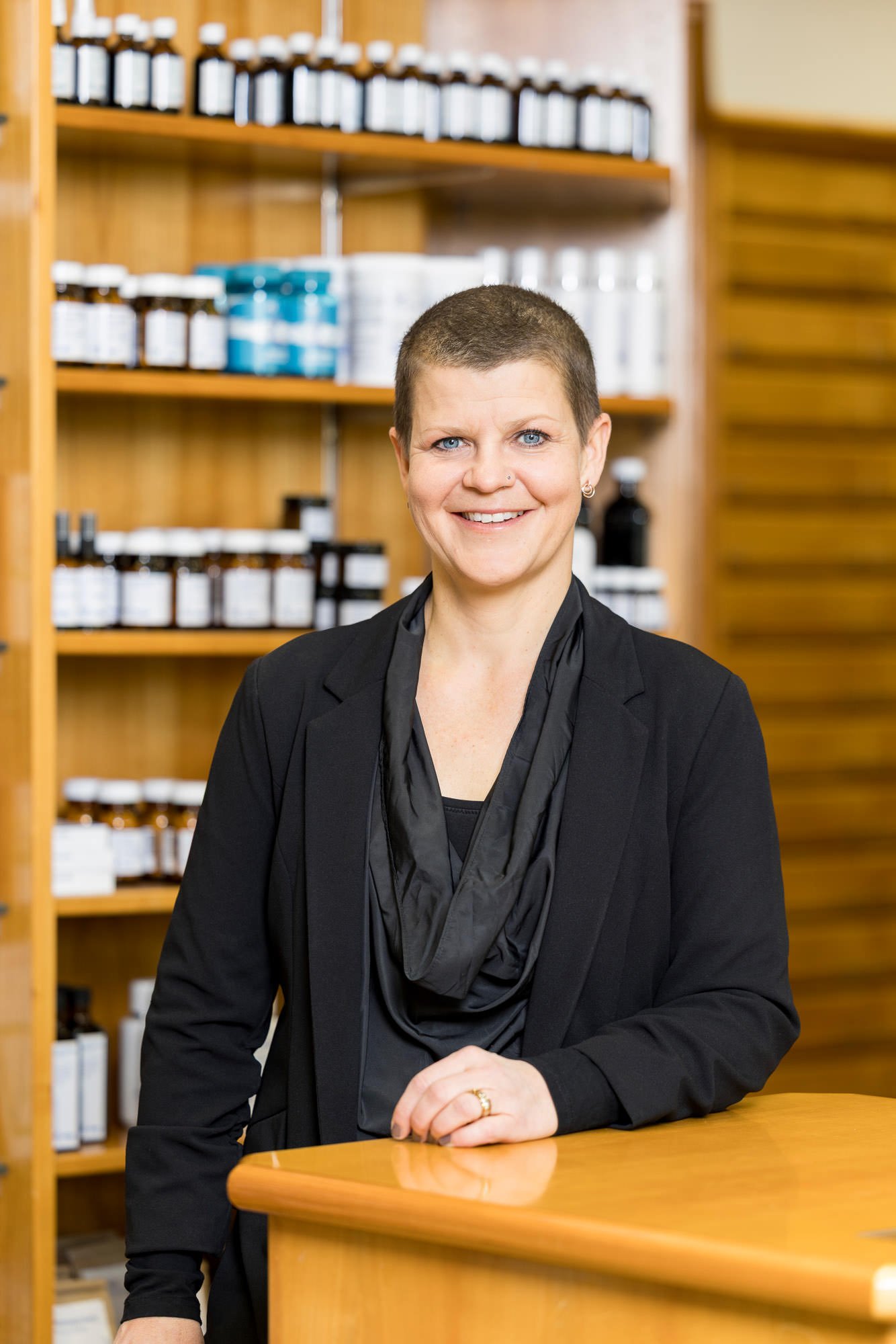 Eine lächelnde Frau mit kurzen Haaren steht an einem Holztresen in einem Geschäft für natürliche Produkte. Im Hintergrund sind Regale mit verschiedenen Flaschen und Behältern zu sehen, die Pflege- und Gesundheitsprodukte enthalten.