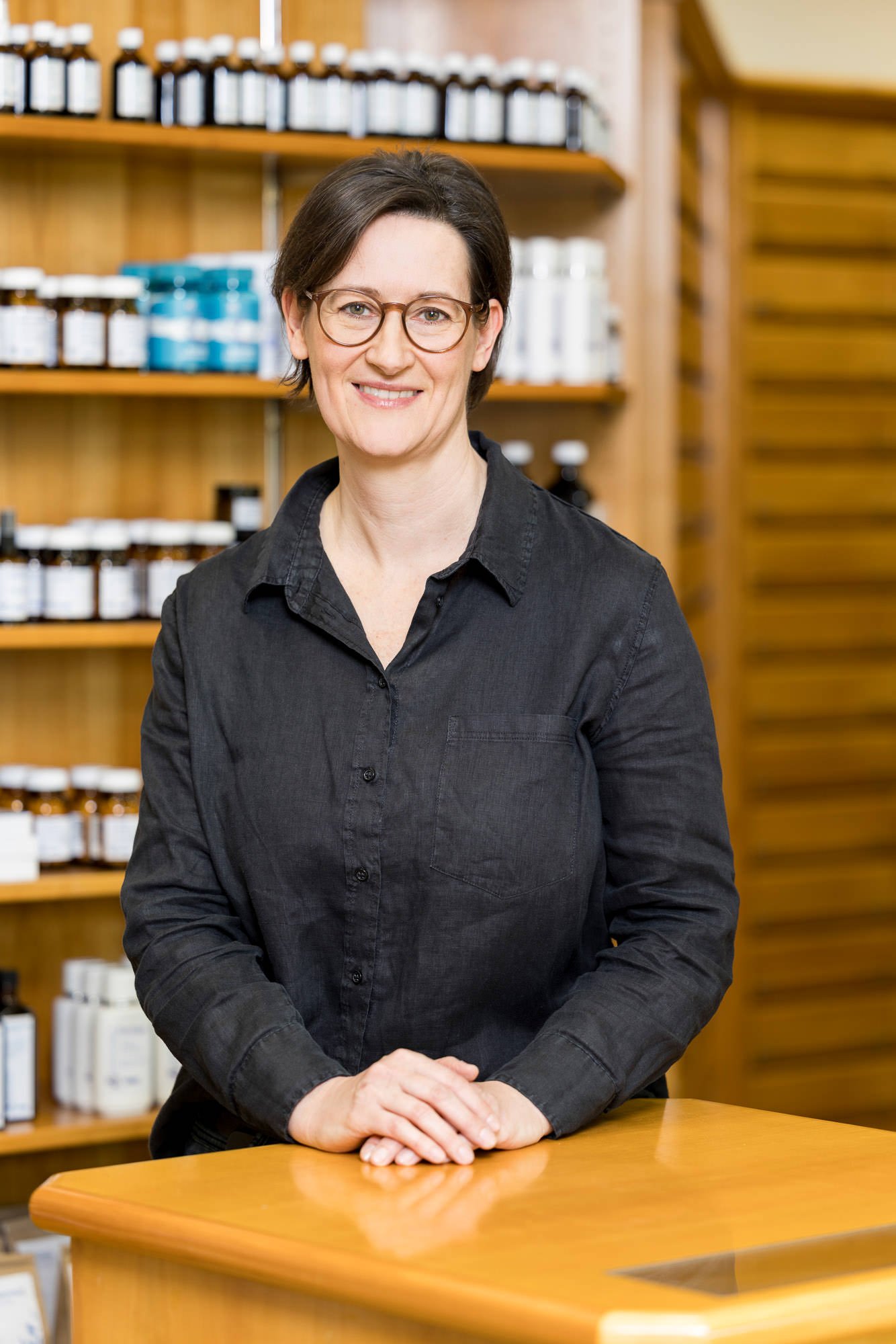 Eine Frau mit Brille steht an einem Empfangstresen in einem Geschäft, umgeben von Regalen mit verschiedenen Fläschchen und Produkten. Sie trägt ein schwarzes Hemd und lächelt freundlich.