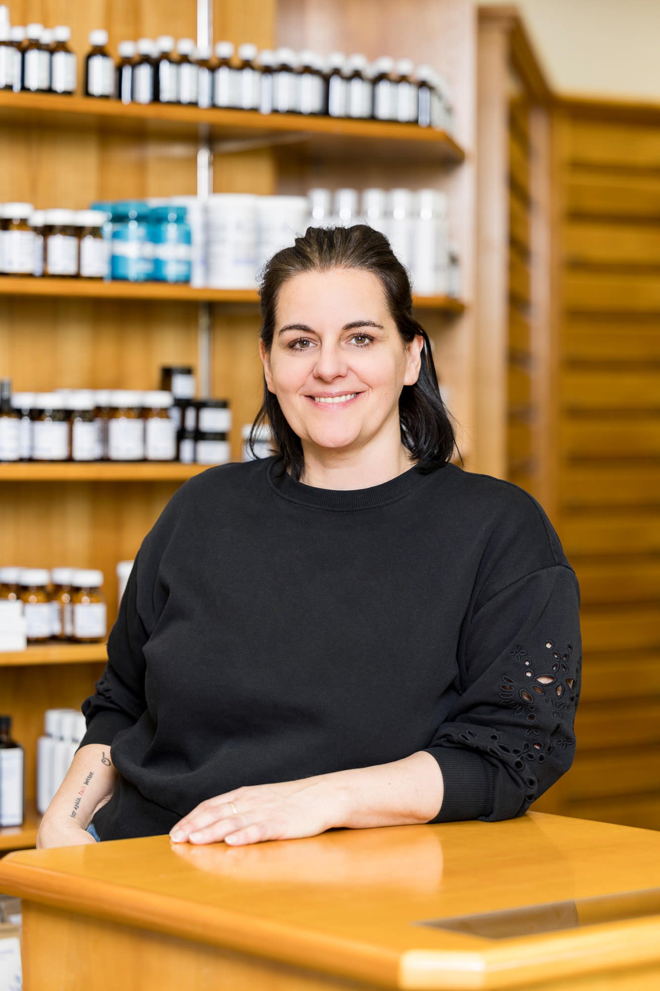 Porträt einer Frau in einem schwarzen Sweatshirt, die an einem Holztisch in einer Apotheke sitzt. Im Hintergrund sind Regale mit verschiedenen Fläschchen und Produkten zu sehen. Die Frau lächelt freundlich und hat ihre Hände auf dem Tisch abgelegt.