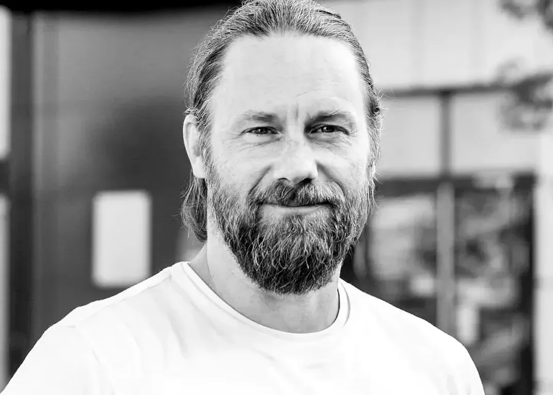 Schwarz-weiß Porträt eines Mannes mit einem Bart, der ein lässiges T-Shirt trägt. Der Mann hat lange Haare, die zu einem Zopf gebunden sind, und lächelt freundlich. Im Hintergrund sind unscharfe Gebäude sichtbar.