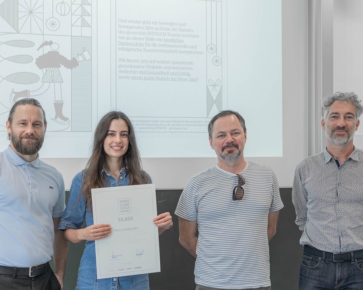 Eine Gruppe von vier Personen posiert für ein Foto bei einer Preisverleihung. Eine junge Frau hält ein Zertifikat in der Mitte, während die anderen drei Männer neben ihr stehen. Die Umgebung ist hell erleuchtet und zeigt eine Präsentation im Hintergrund.