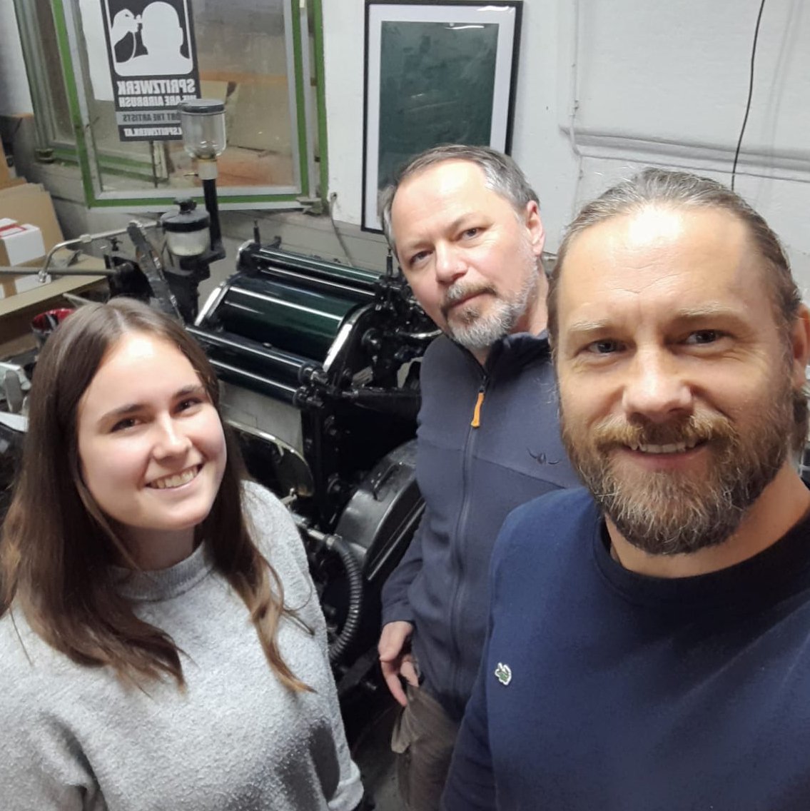 Drei Personen in einer Druckerei, vor einer alten Druckmaschine. Die Gruppe besteht aus einer jungen Frau mit langen Haaren, einem Mann in einem grauen Pullover und einem weiteren Mann in einem schwarzen Pullover. Im Hintergrund sind verschiedene Druckmaterialien und eine grüne Fensterbank zu sehen.