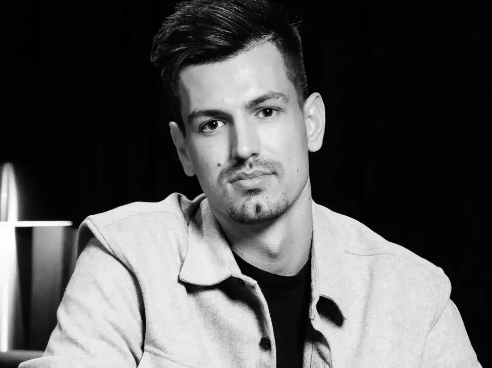 Porträt eines jungen Mannes mit kurzem Haar und gepflegtem Bart, der in einem hellen Oberteil posiert. Der Hintergrund ist dunkel, was einen Kontrast zu seiner hellen Kleidung schafft. Die Stimmung des Bildes ist seriös und ansprechend.