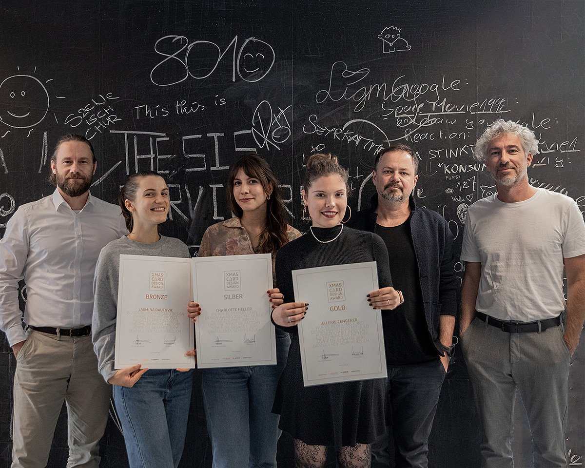 Gruppe von sechs Personen, darunter drei Frauen und drei Männer, die stolz ihre Zertifikate in einer kreativen Umgebung mit einer schwarzen Wand voller Kritzeleien und Text präsentieren. Die Frauen halten ihre Auszeichnungen in der Mitte, während die Männer an den Seiten stehen.