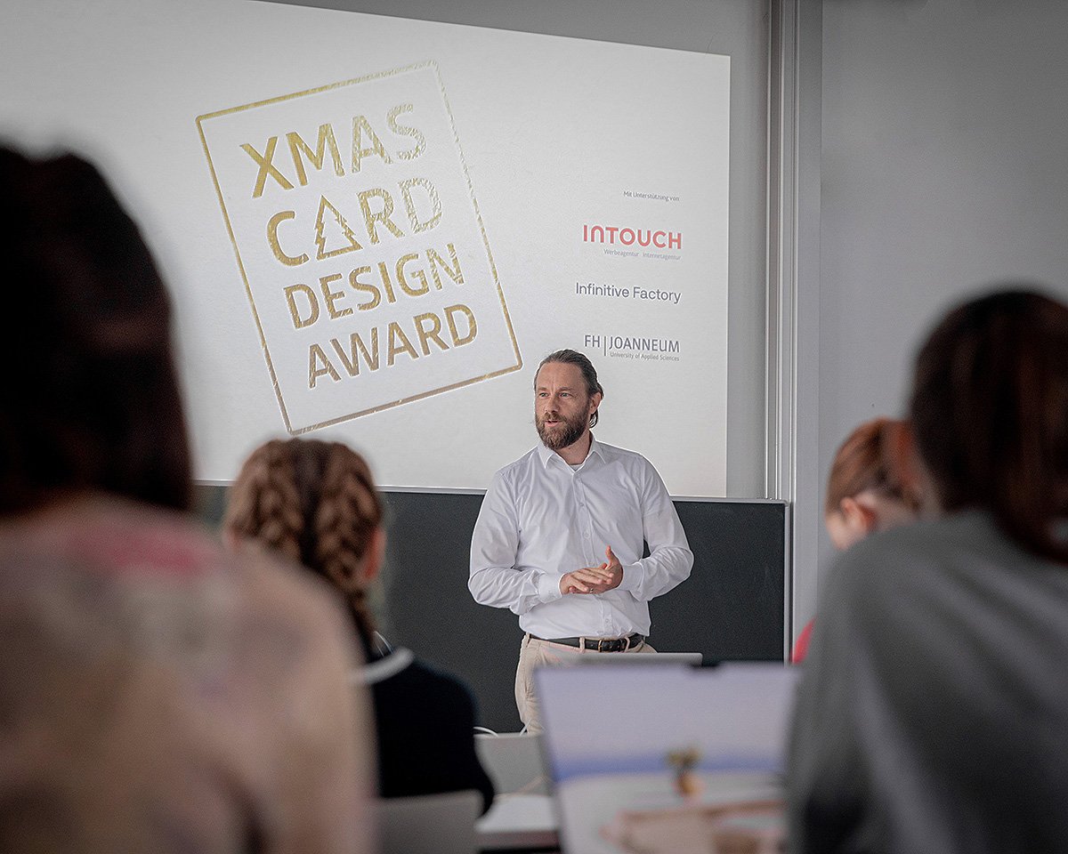 Ein Mann präsentiert während einer Veranstaltung den XMAS CARD DESIGN AWARD. Im Hintergrund ist das Logo des Awards sowie die Sponsoren INT TOUCH, Initiative Factory und ZHI DANIELM zu sehen. Die Zuhörer sind teilweise im Bild, aufmerksam auf die Präsentation gerichtet.