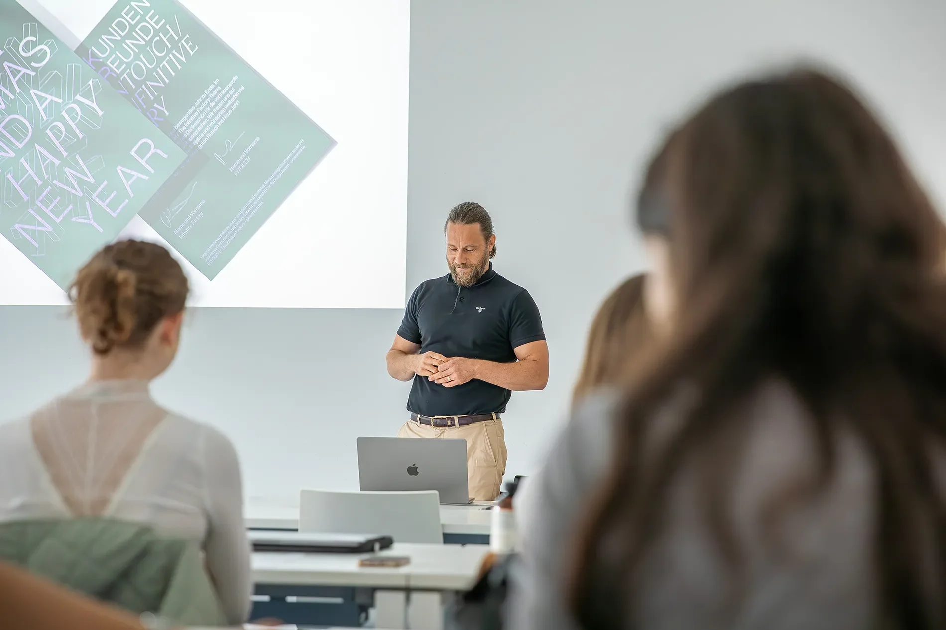 Ein Dozent steht vor einer Gruppe von Studierenden in einem modernen Klassenraum. Auf einer Leinwand im Hintergrund sind Grafiken zu sehen, die mit Neujahrsgrüßen in Verbindung stehen. Der Dozent trägt ein schwarzes Polohemd und beige Hosen, während die Studierenden aufmerksam zuhören.