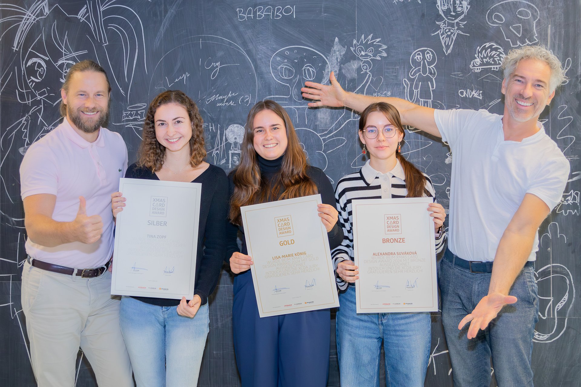 Gruppe von vier Personen, die stolz ihre Zertifikate in einem kreativen Umfeld halten, vor einer Wand mit bunten Zeichnungen. Zwei Männer, einer mit Daumen hoch, und zwei Frauen, die in die Kamera lächeln.