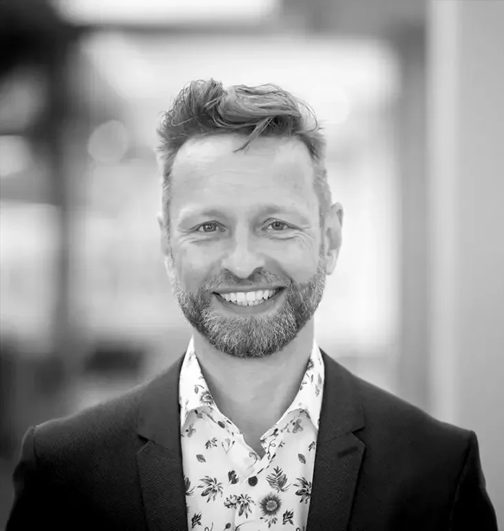 Professionelles Porträt eines Mannes mit kurzem, welligem Haar und Bart, der ein lächelndes Gesicht zeigt. Er trägt einen schwarzen Anzug und ein helles, blumiges Hemd. Der Hintergrund ist unscharf und vermittelt eine moderne Atmosphäre.