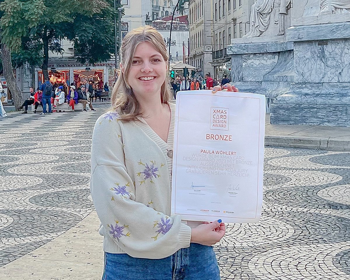 Junge Frau mit blonden Haaren hält ein Zertifikat in der Hand, das den Bronze-Preis für einen Wettbewerb zeigt. Sie steht auf einem Platz mit Steinmuster, umgeben von Menschen und historischen Gebäuden.