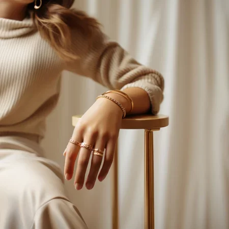Eine elegante Hand mit mehreren goldenen Ringen und Armbändern ruht auf einem modernen, minimalistischen Beistelltisch. Die Person trägt einen beigen Pullover und befindet sich in einem hellen, stilvollen Raum mit sanften Vorhängen im Hintergrund.