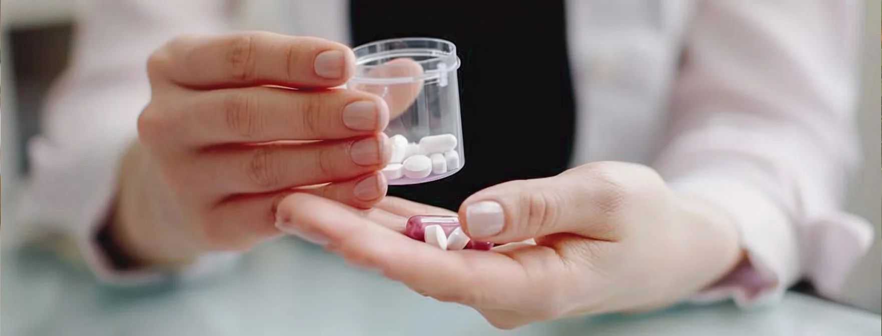 Eine Person hält ein transparentes Behältnis mit Medikamenten in einer Hand und lässt einige Tabletten in die andere Hand fallen. Die Tabletten sind in verschiedenen Farben und Formen und befinden sich auf einem Tisch.