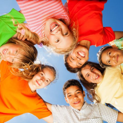 Gruppe von fröhlichen Kindern, die im Freien stehen und in die Kamera lächeln. Sie tragen bunte T-Shirts und bilden einen Kreis, während sie sich an den Händen halten. Der Himmel ist blau und strahlend.
