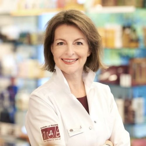 Eine Frau in einem weißen Laborkittel lächelt freundlich. Sie steht vor einem farbenfrohen Hintergrund mit verschiedenen Produkten. Die Atmosphäre vermittelt Professionalität und Fachkompetenz.