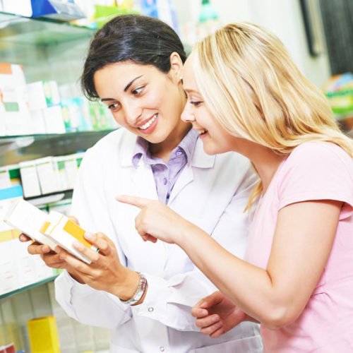 Eine Pharmazeutin berät eine Kundin in einer Apotheke. Beide schauen auf ein Produkt, während die Pharmazeutin Informationen dazu gibt. Der Hintergrund zeigt Regale mit verschiedenen Gesundheitsprodukten und Arzneimitteln.