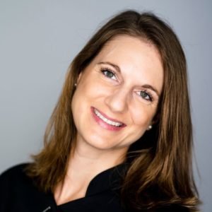 Ein Porträtfoto einer lächelnden Frau mit langen, braunen Haaren. Sie trägt ein schwarzes Oberteil und hat einen freundlichen Gesichtsausdruck vor einem neutralen grauen Hintergrund.