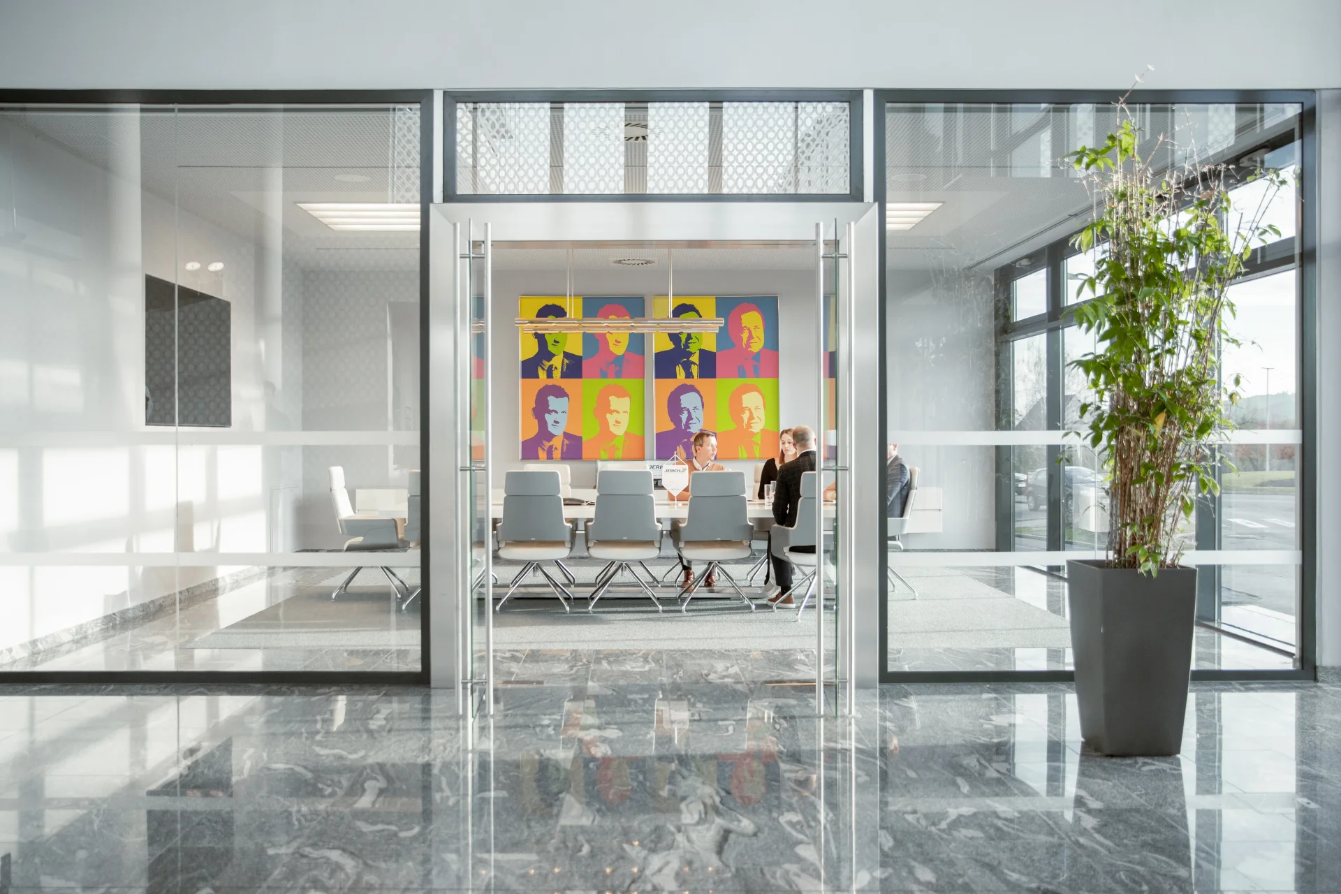 Moderne Büroumgebung mit Glaswänden, eleganten Konferenzstühlen und einem bunten Wandbild. Der Raum ist hell und geräumig, mit einem großen Fenster, das Tageslicht hereinlässt. Eine Pflanze im Topf ergänzt die Einrichtung.