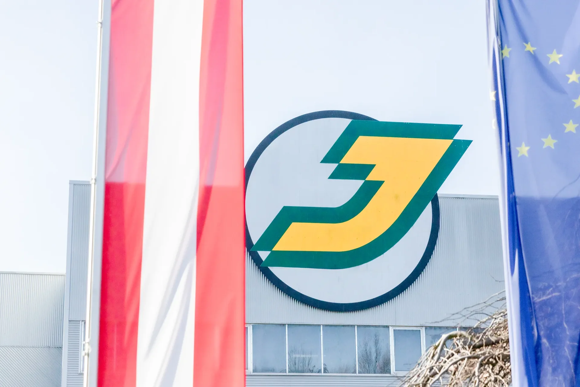 Logo eines Unternehmens mit grünen und gelben Elementen, umgeben von Flaggen, darunter die Flagge Österreichs und die EU-Flagge.