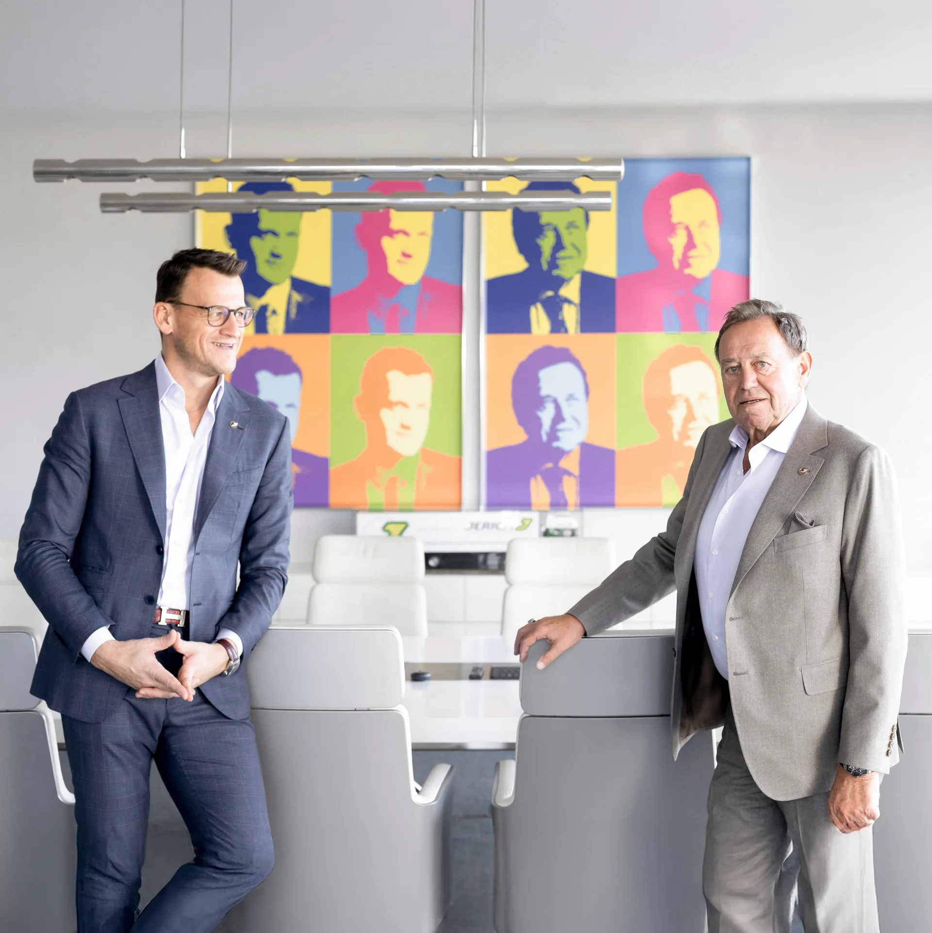 Two businesspeople in a modern conference room stand in front of a colorful work of art featuring several portraits. The man on the left wears a dark suit and glasses, while the man on the right is dressed in a light-colored suit. The conference room has minimalist furnishings and exudes professionalism.