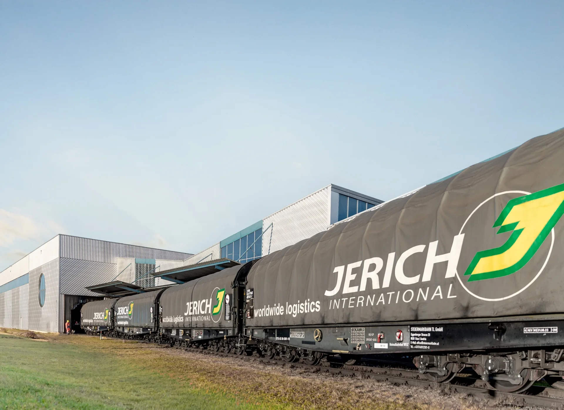 Zug mit Güterwagen der Firma Jerich International vor einem modernen Gebäude, das auf weltweite Logistik spezialisiert ist.