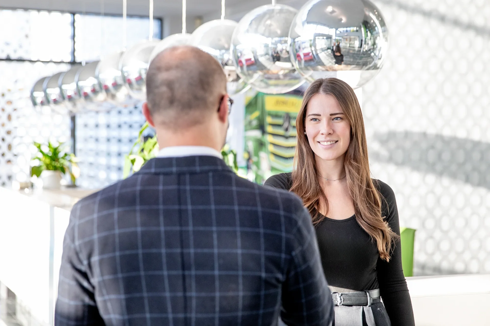 Ein Gespräch zwischen zwei Personen in einem modernen Büro. Eine Frau mit langen, braunen Haaren lächelt, während sie einem Mann, der ihr den Rücken zuwendet, zuhört. Im Hintergrund sind dekorative, glänzende Kugeln und Pflanzen zu sehen, die das zeitgenössische Ambiente des Raumes unterstreichen.