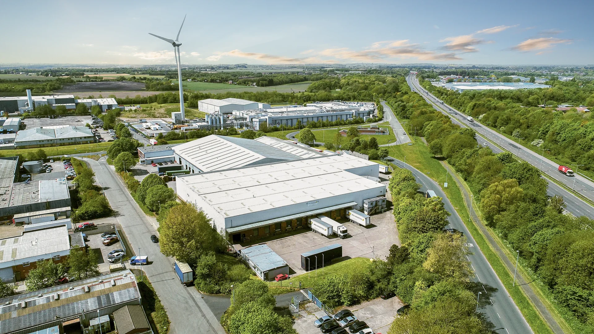 Aerial view of an industrial area with several warehouses and a wind turbine in the background. The scene shows a well-developed road with parked trucks and surrounding green areas. The sky is clear with a few clouds.