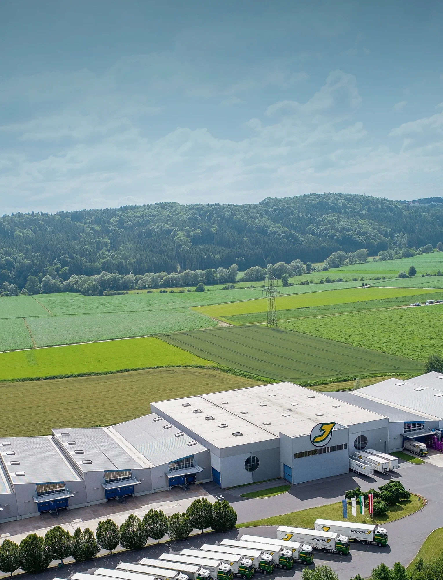 Luftaufnahme eines modernen Lagerhauses mit grünen Feldern im Hintergrund, umgeben von Hügeln und bewaldeten Flächen. Die Gebäude sind mit Lkw und einer klaren blauen Himmelkulisse zu sehen.