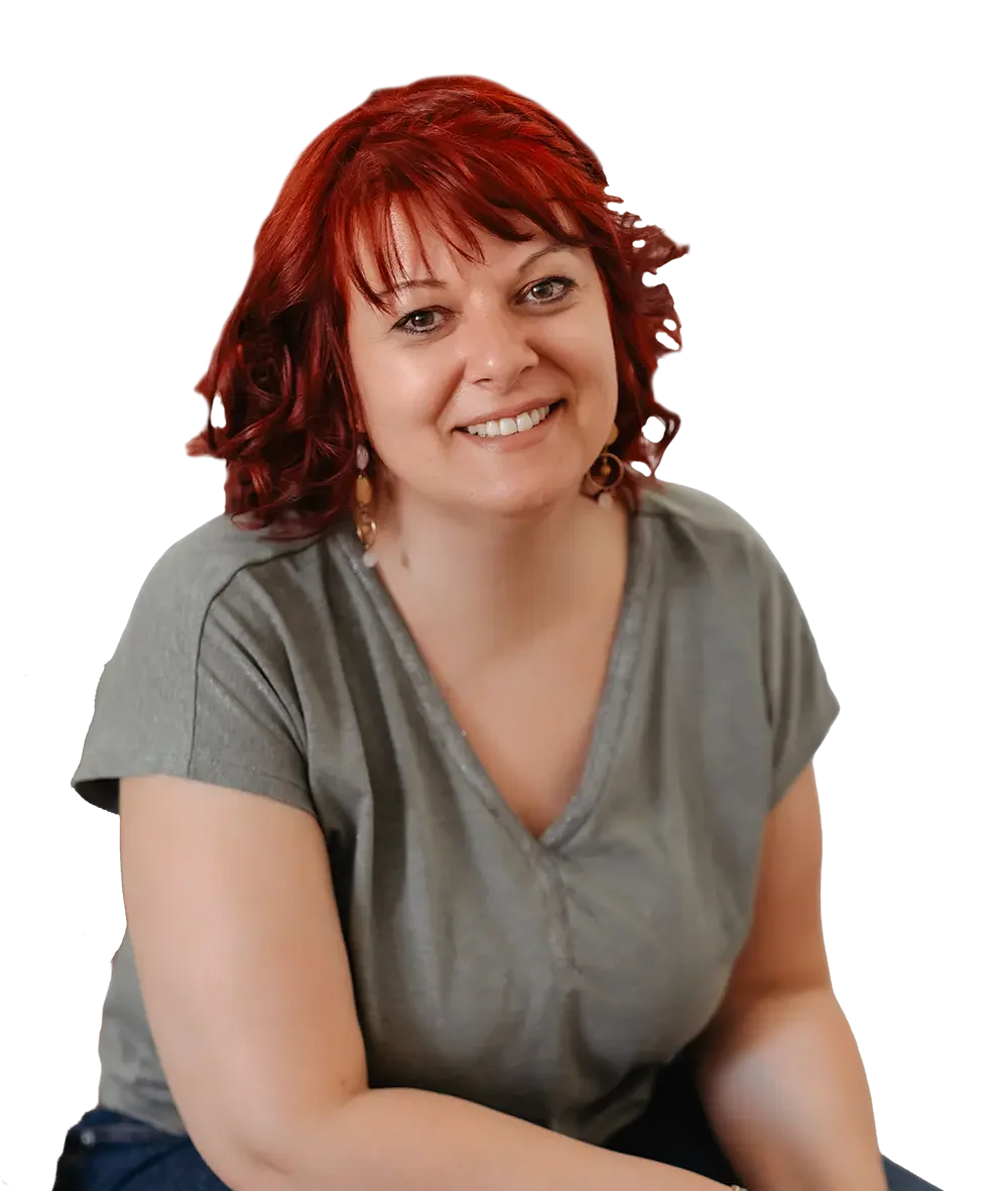 Porträt einer lächelnden Frau mit roten, lockigen Haaren, die ein graues T-Shirt trägt. Sie sitzt entspannt und blickt direkt in die Kamera. Die Aufnahme hat einen neutralen Hintergrund und vermittelt eine freundliche, einladende Atmosphäre.