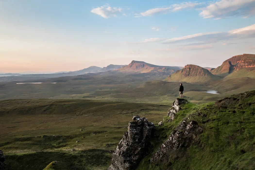 Ein Wanderer steht auf einem Felsen und blickt über die weite, grüne Landschaft der Isle of Skye in Schottland. Die sanften Hügel und markanten Berge sind im Hintergrund sichtbar, während der Himmel von sanften Farben des Sonnenuntergangs beleuchtet wird.