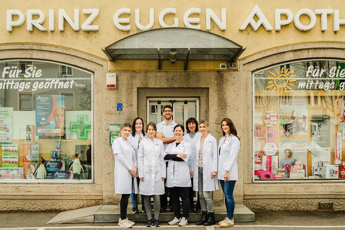 Gruppe von acht Apothekern in weißen Laborkitteln vor der Prinz Eugen Apotheke. Die Apotheke zeigt ein breites Sortiment an Gesundheitsprodukten in den Fenstern. Freundliche Atmosphäre und professionelle Präsentation.