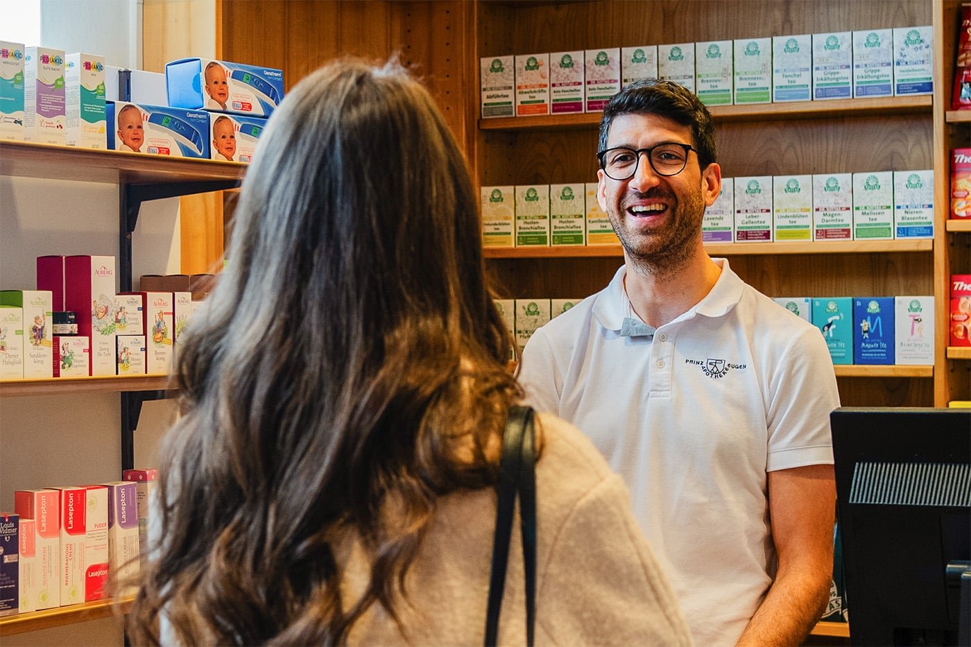 Ein lächelnder Mann in einem weißen Polohemd steht hinter einer Theke in einer Apotheke, während er mit einer Frau spricht, die ihm den Rücken zukehrt. Im Hintergrund sind Regale mit verschiedenen pharmazeutischen Produkten sichtbar.