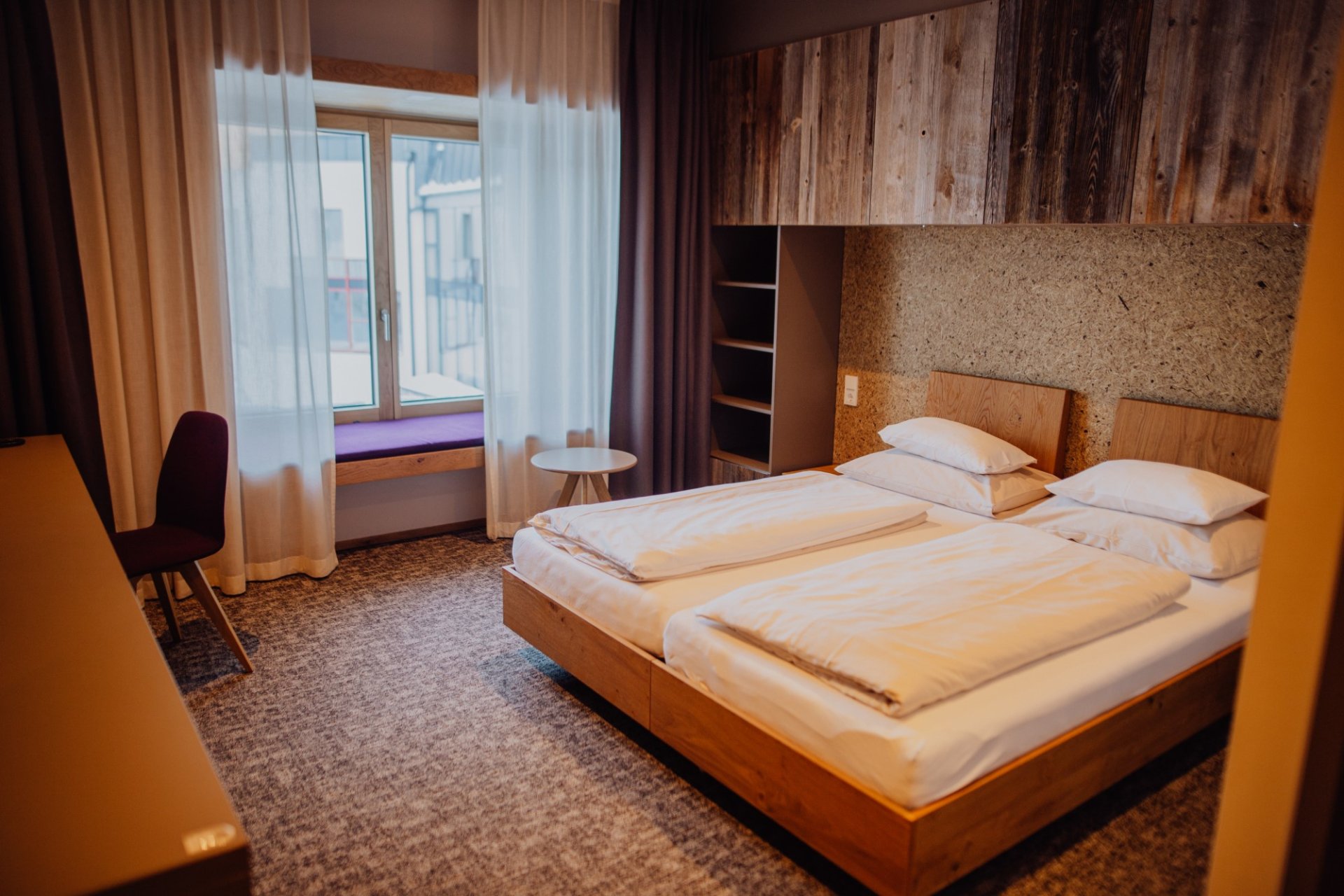 Modernes Hotelzimmer mit zwei Einzelbetten, Holz- und Textiloberflächen, großen Fenstern mit Vorhängen, einem kleinen Tisch und einem Stuhl, ideal für Erholung und Entspannung.