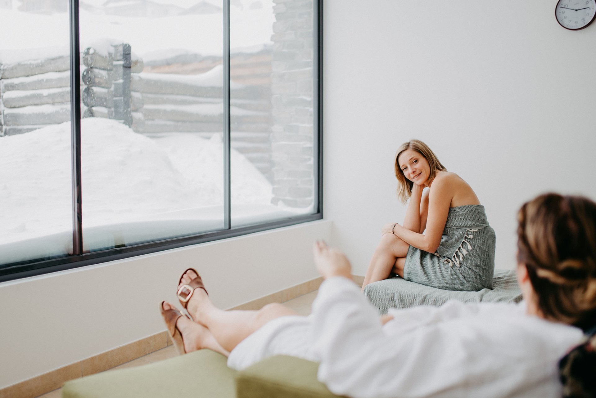 Zwei Frauen in einem modernen Raum mit großen Fenstern, die auf eine schneebedeckte Landschaft blicken. Eine Frau sitzt auf einer Liege und trägt ein graues Kleid, während die andere in einem Bademantel auf einer anderen Liege liegt. Die Atmosphäre ist entspannend und lädt zur Erholung ein.