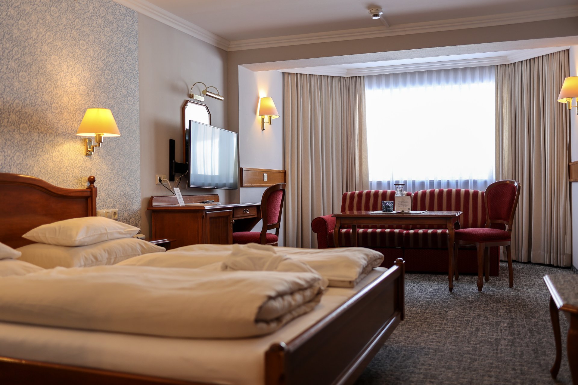 Ein modernes Hotelzimmer mit einem Doppelbett, zwei Kissen und einem Holzkopfteil. Im Hintergrund ist ein Fernseher an der Wand montiert. Vor dem Fenster befinden sich helle Vorhänge, die Tageslicht hereinlassen. Eine Sitzecke mit einem roten Sofa und einem Tisch ist ebenfalls sichtbar, zusammen mit einem Holzschreibtisch und einem Stuhl.
