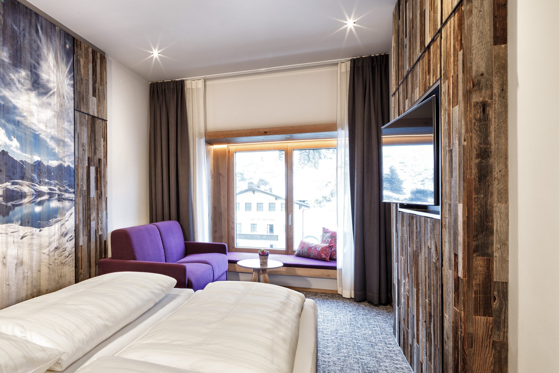 Helles Hotelzimmer mit einem modernen Design, ausgestattet mit zwei Einzelbetten, einem lila Sofa und einem Fernseher. Große Fenster bieten einen Blick auf die Außenansicht. Die Wände sind teilweise mit Holz verkleidet, was eine warme Atmosphäre schafft.