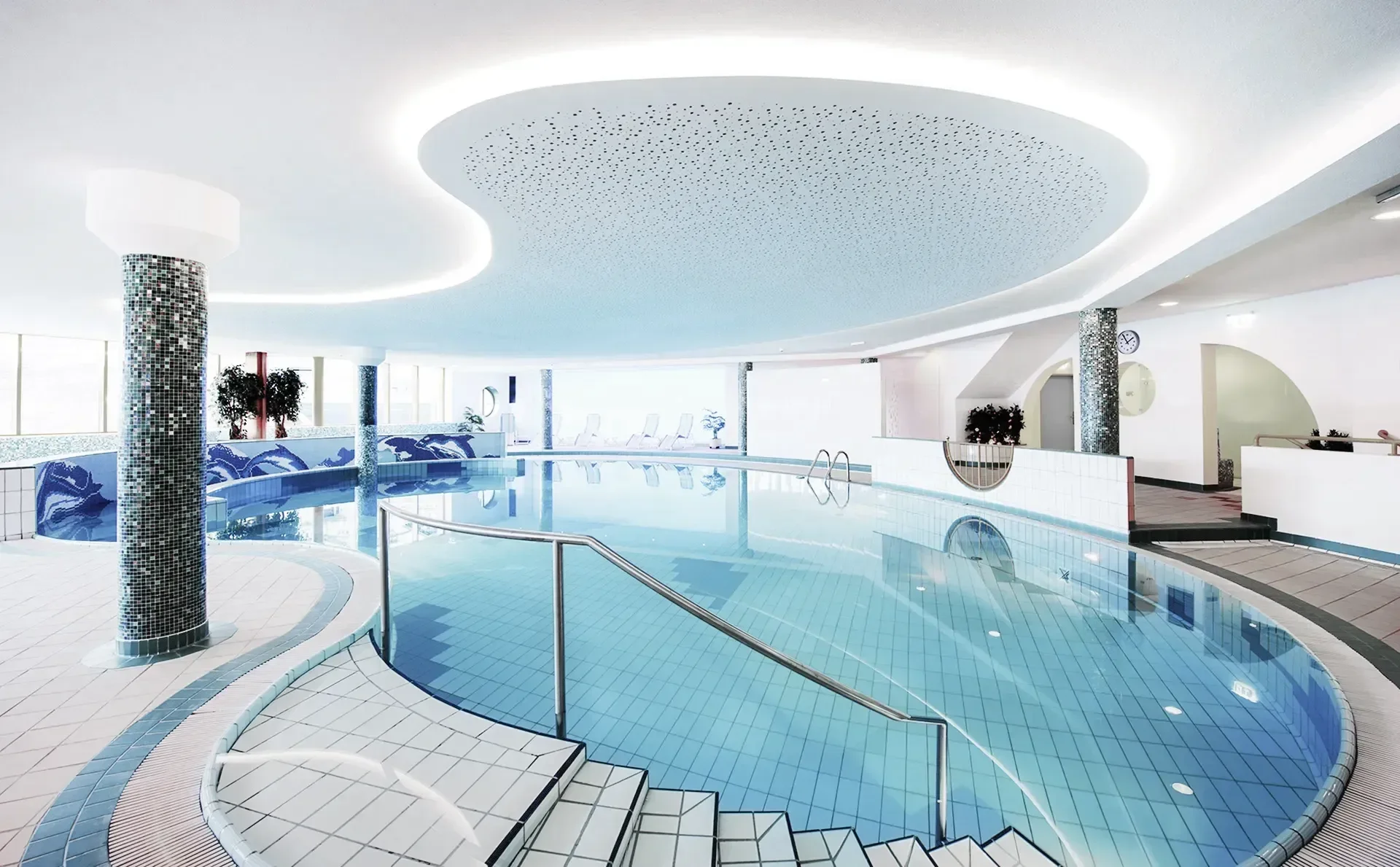 Moderne Innenansicht eines Schwimmbereichs mit einem stilvollen Pool, umgeben von eleganten Fliesen und einem hellen, einladenden Ambiente. Die Decke ist mit dekorativen Lichtern gestaltet und große Fenster bieten viel Tageslicht.