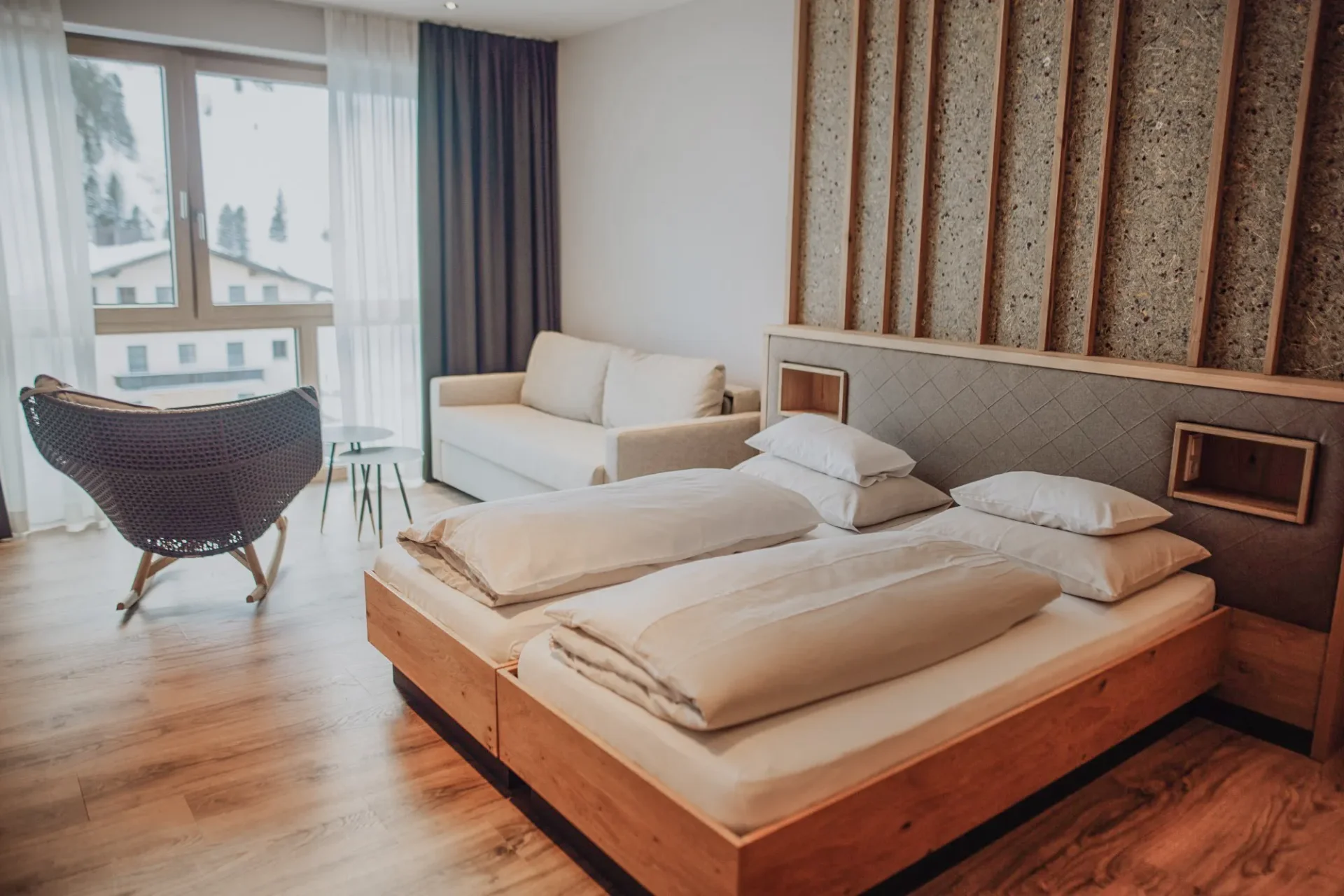 Bright, modern bedroom with two beds, comfortable pillows and a cozy seating area. Large windows provide plenty of natural light and views of the surrounding area. The furnishings combine wood with neutral colors for a relaxed atmosphere.