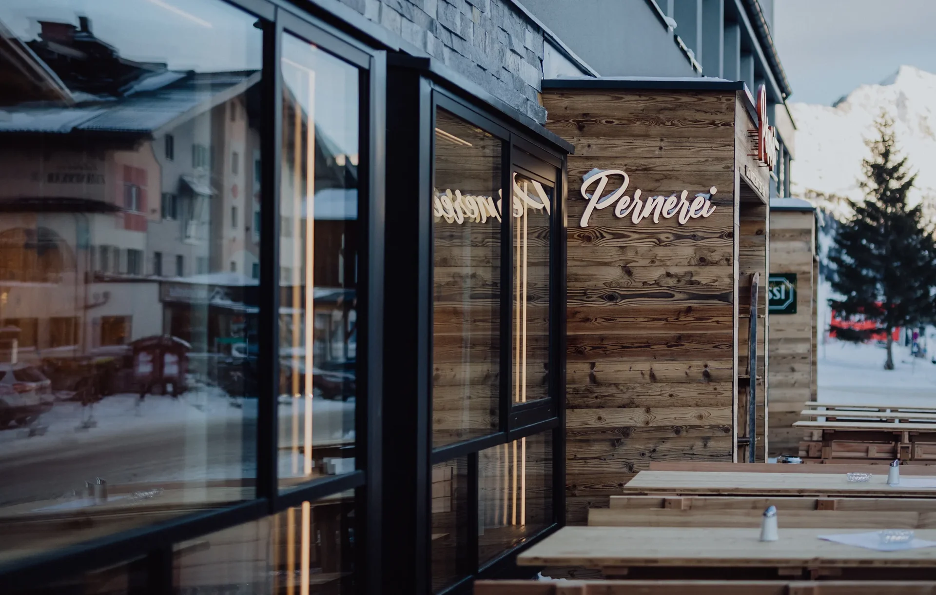 Ansicht eines modernen Restaurants mit Holzverkleidung und großen Fenstern, das den Namen 'Pernerei' trägt, in einer schneebedeckten Umgebung.