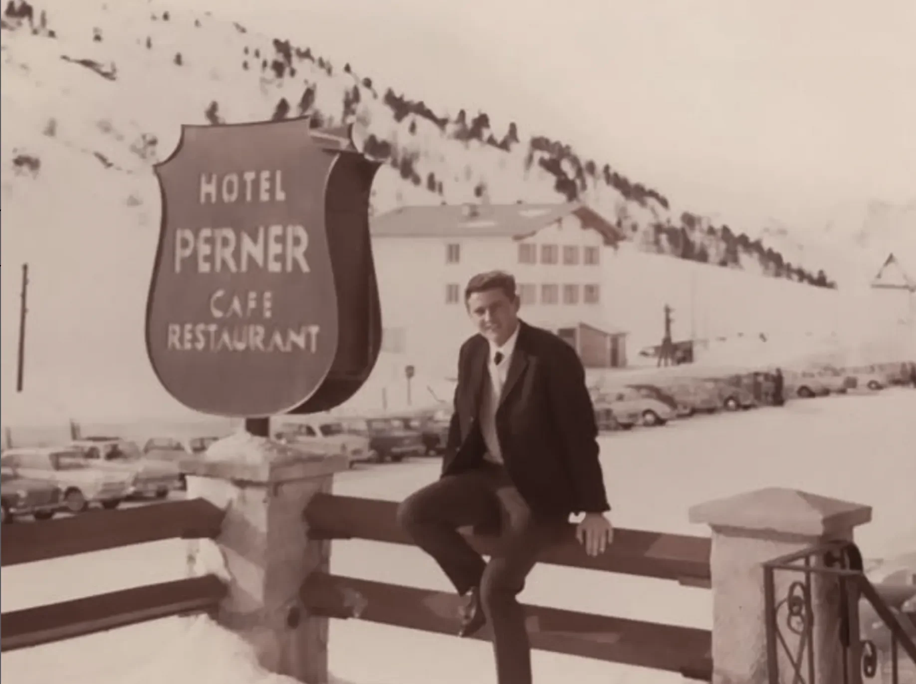 Ein Mann in formeller Kleidung sitzt auf einer Brüstung vor einem Schild des Hotel Perner, das ein Café und Restaurant anzeigt. Im Hintergrund sind schneebedeckte Berge und ein geparkter Autos in einer Winterlandschaft zu sehen.