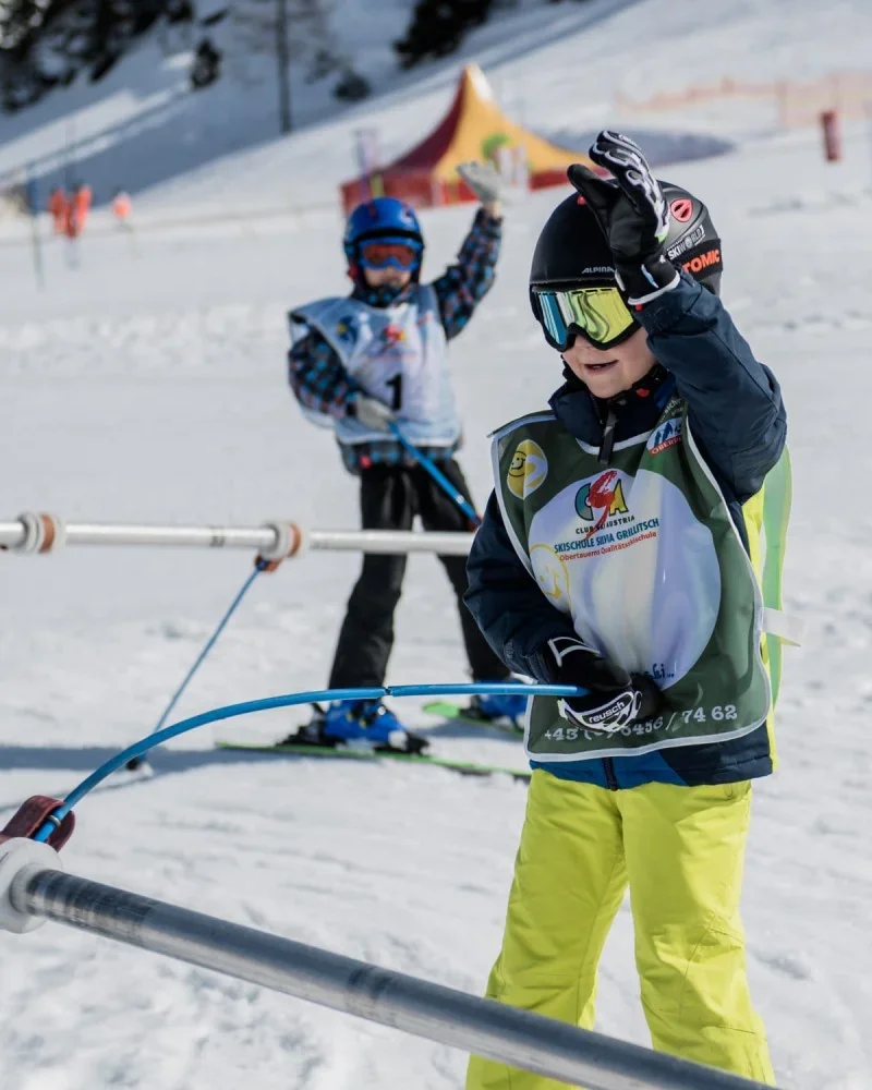 Zwei Kinder in Skiausrüstung stehen auf einer Skipiste. Das vordere Kind trägt eine schwarze Mütze und gelbe Skihose, während das andere Kind im Hintergrund einen blauen Helm und eine Jacke trägt. Beide Kinder scheinen an einem Skikurs teilzunehmen, während sie an einem Skilehrer oder einer Übung teilnehmen.