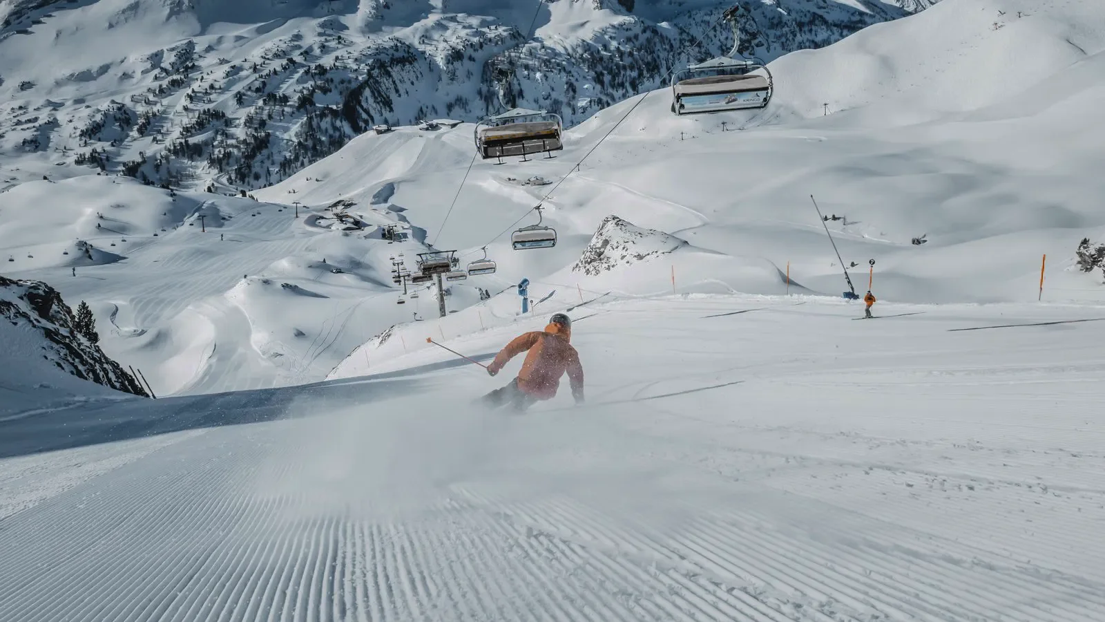 Ein Skifahrer fährt auf einer schneebedeckten Piste, umgeben von Bergen und Skiliften. Die frisch präparierte Piste zeigt klare Spuren im Schnee, während der Himmel blau ist.