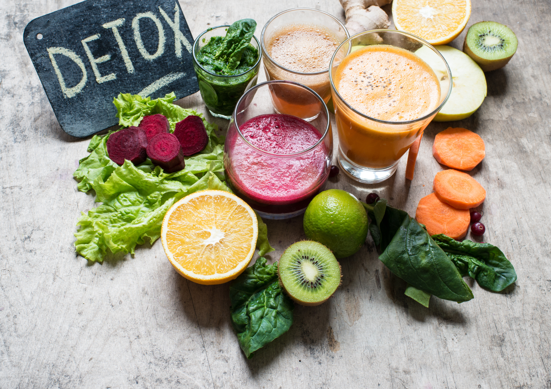 Eine Auswahl an frischen Säften und Obst für eine Detox-Diät, einschließlich grünem Blattgemüse, Rüben, Orangen, Limetten, Kiwi, Karotten und einem kleinen Tafelschild mit der Aufschrift 'DETOX'.