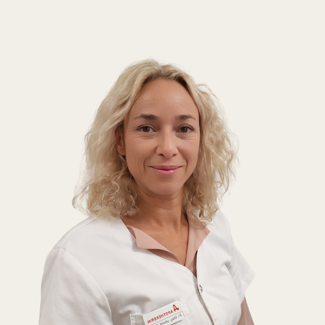 Porträt einer Frau mit lockigem, blondem Haar, die in einem weißen Kittel gekleidet ist. Sie lächelt und steht vor einem neutralen Hintergrund. Das Bild vermittelt Professionalität und Freundlichkeit.