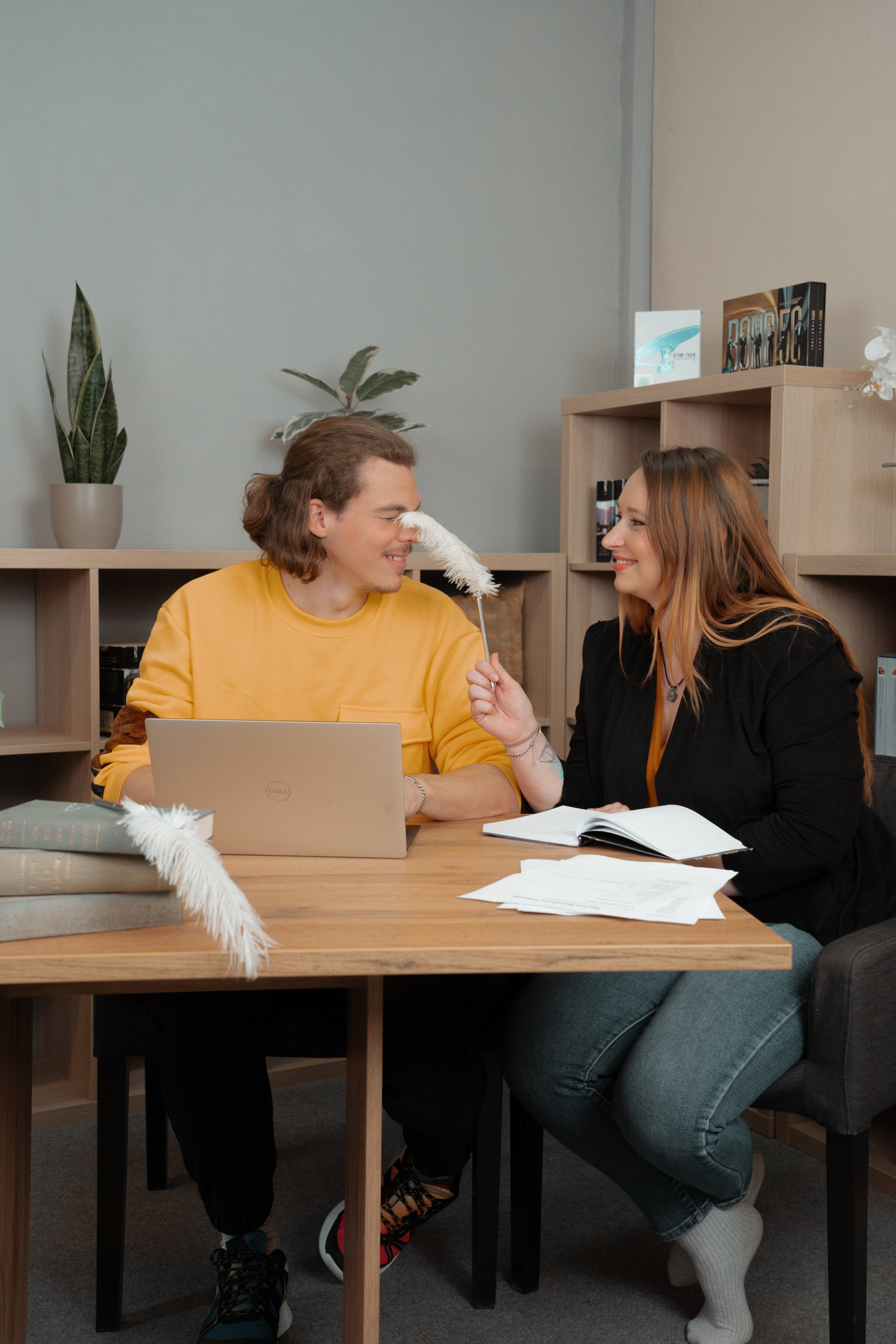 Ein junger Mann in einem gelben Pullover sitzt an einem Tisch mit einem Laptop, während eine junge Frau in einem schwarzen Blazer ihm mit einer Feder ins Gesicht tippt. Auf dem Tisch liegen einige Blätter Papier und ein Buch. Im Hintergrund sind Pflanzen und Regale zu sehen.