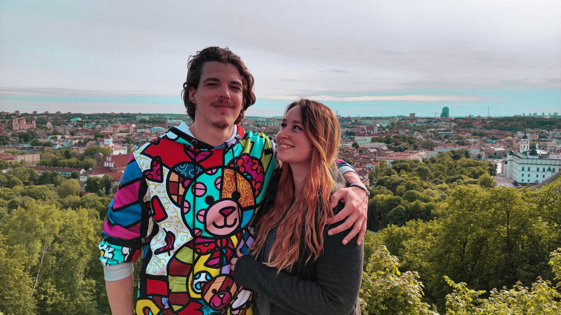 Junge und Mädchen posieren vor einer Stadtlandschaft, umgeben von Bäumen, mit einer bunten Hoodie-Jacke und lächelndem Gesichtsausdruck.