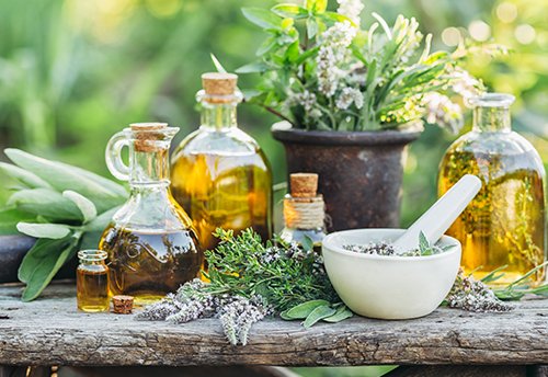 Eine rustikale Anordnung von verschiedenen Flaschen mit Ölen und frischen Kräutern auf einem Holztisch. Im Vordergrund steht ein Mörser mit Kräutern, umgeben von weiteren Pflanzen und Flaschen, die in einem Gartenumfeld platziert sind.