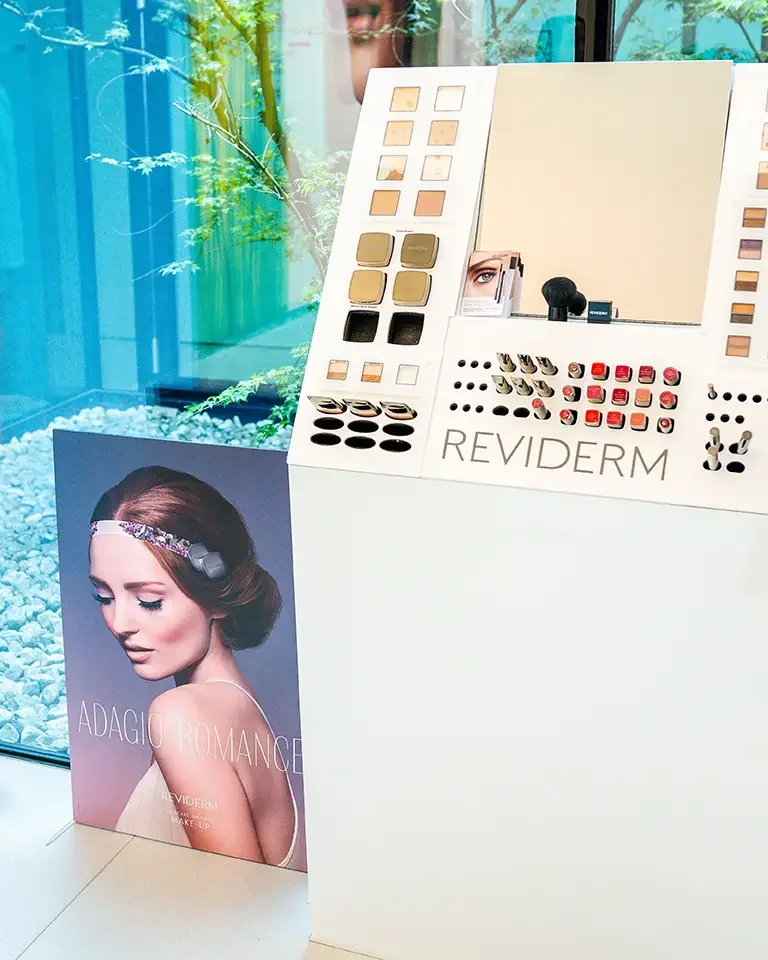 Schönheitsstand mit Spiegel und Kosmetikprodukten, präsentiert von Reviderm, vor einem Werbeplakat mit dem Titel "Adagio Romance". Der Bereich ist hell erleuchtet und bietet eine Vielzahl von Farbtönen für Make-up und Pflege.